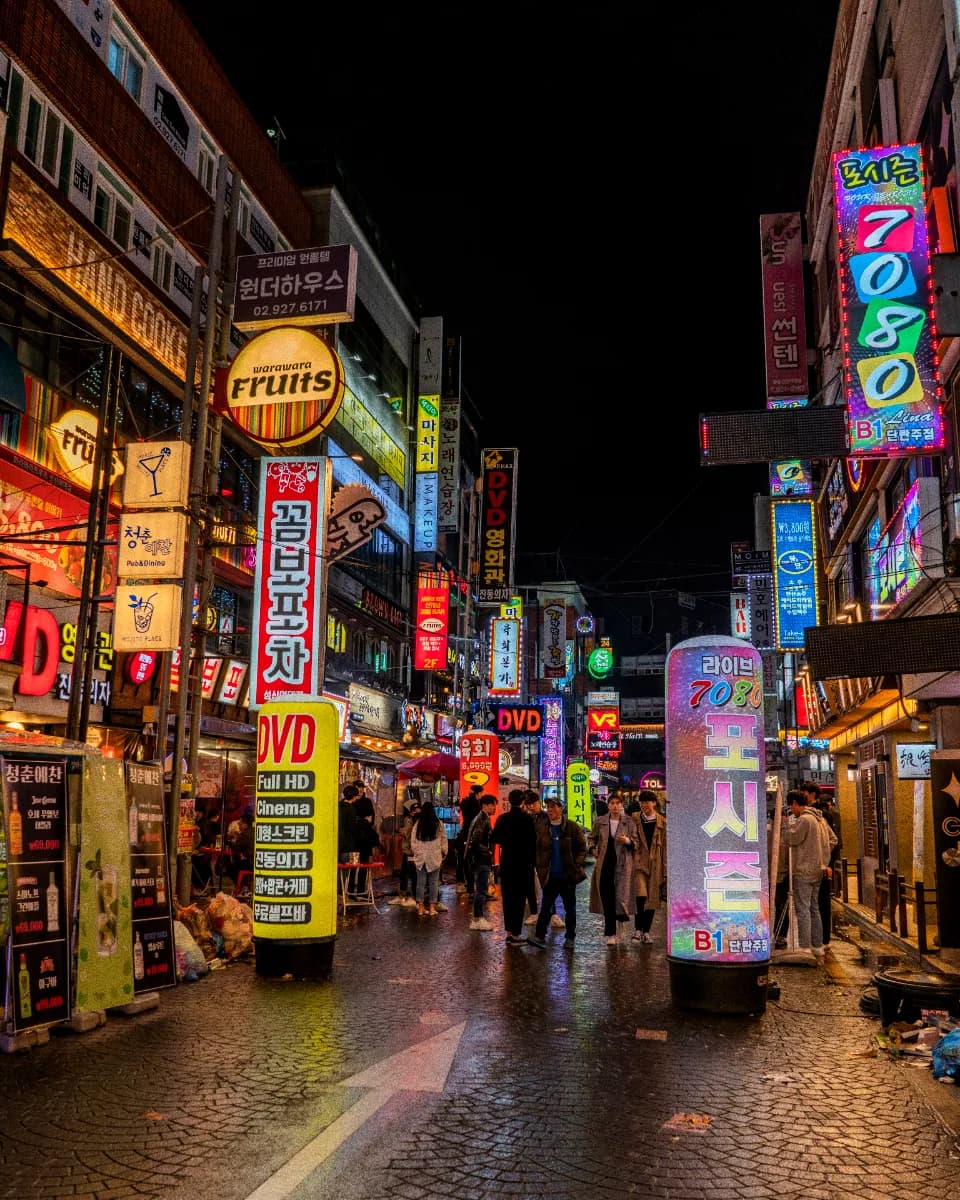 Layered street signs and a busy pavement crowd under coloured light